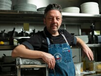A man wearing a blue denim apron stands with his right arm resting on metal pasta maker. Behind him are metal shelves containing different plates and utensils. 