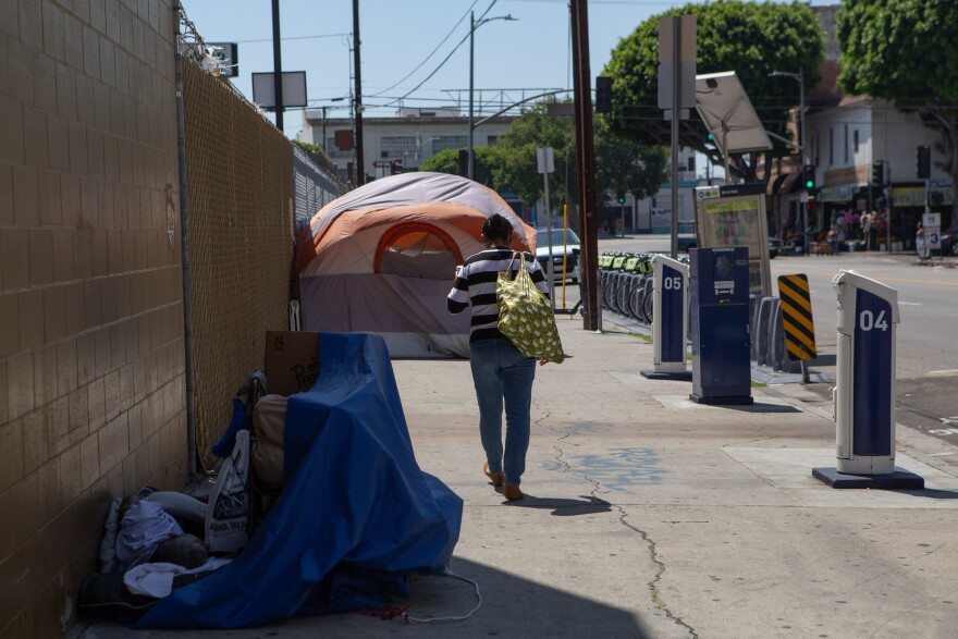 A person walks down a street toward a tent on the sidewalk. Personal belongings partially covered by a blue tarp are also on the sidewalk.