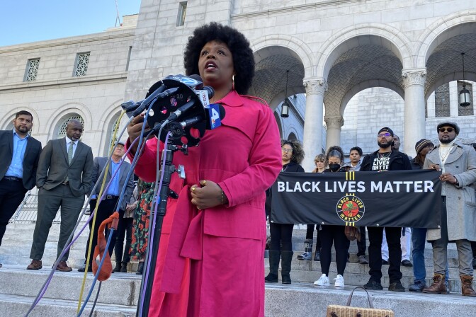 BLM co-founder Patrisse Cullors speaks into several microphones outside LA city hall. A banner behind her reads "Black Lives Matter." City council members and several activists stand behind her. Cullors wears a bright pink coat. 
