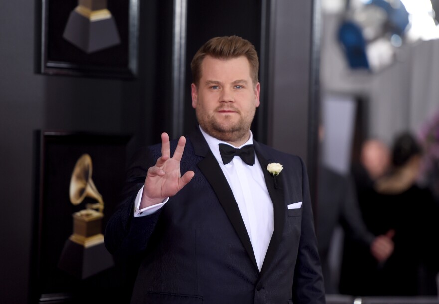 James Corden, dressed in a tuxedo with a yellow boutonnière, raises one hand in a wave. He has light skin and stubble on his chin and cheeks.