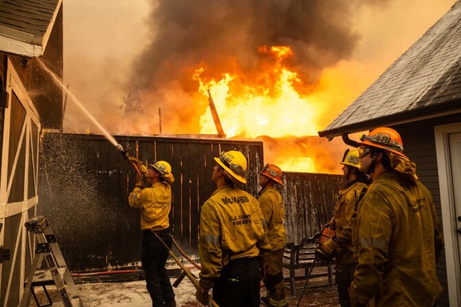 Firefighters spray water onto a home as a building while flames shoot high into the sky in the background, behind a wooden fence.