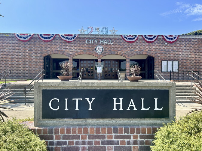 A brick City Hall building features patriotic bunting and a '250' sign, with a stone City Hall sign in front.
