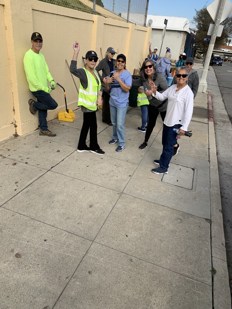 A group of volunteers smiling and waving on a city sidewalk. Several are wearing neon green vests, hats and sunglasses.