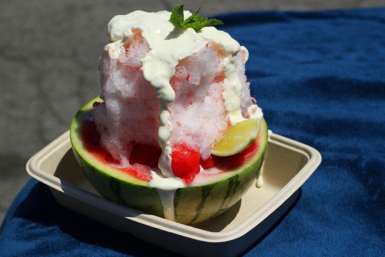 white cream drizzled on a mound of shaved ice with red syrup sitting inside half of a hollowed out small watermelon