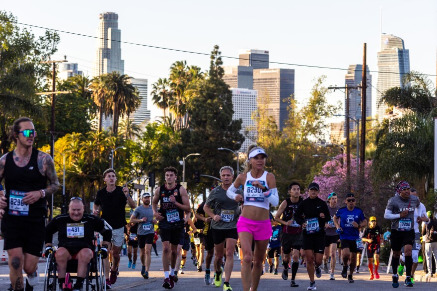 Marathon runners run up a hill with palm trees and downtown L.A. skyscrapers in the background. 
