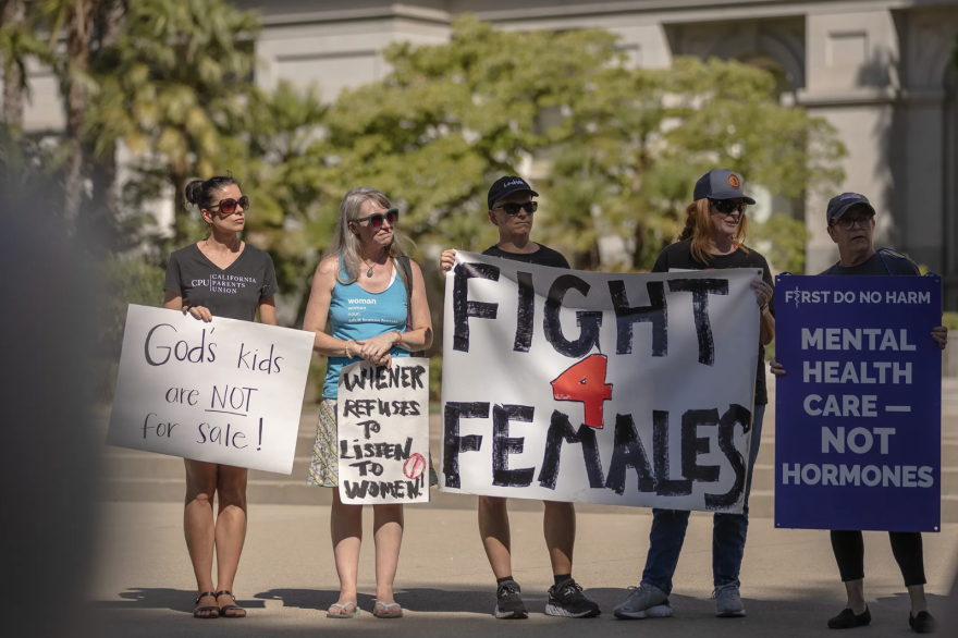 Five people with light skin tone are outside holding protest signs. The biggest sign reads "FIGHT 4 FEMALES" and another reads "God's kids are NOT for sale!"