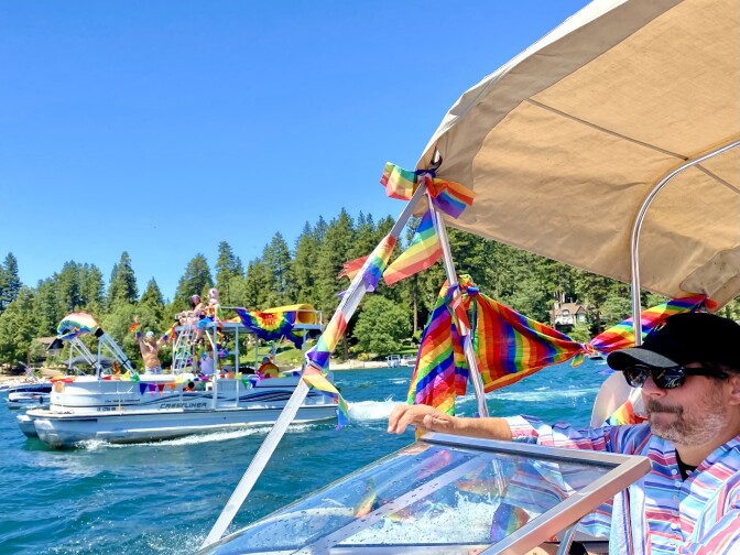 A man with light skin tone and a salt-and-pepper beard with sunglasses  drives a boat festooned with rainbow flags. There is a pontoon boat in the background that also has rainbow flags. Pine trees ring the lake. 