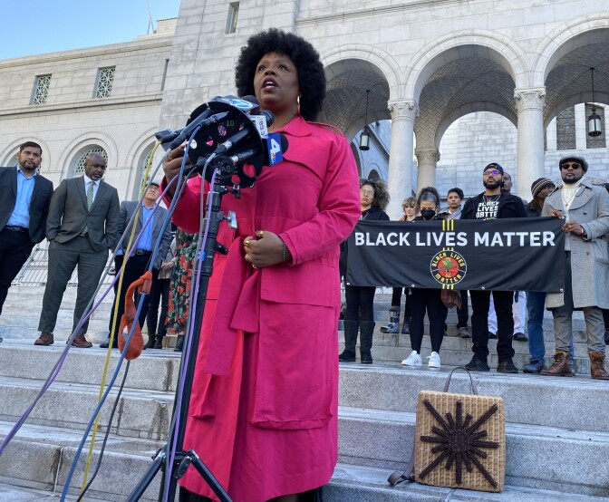 A Black woman wearing a pink long coat speaks into several microphones outside LA city hall. A banner behind her reads "Black Lives Matter." City council members and several activists stand behind her. 