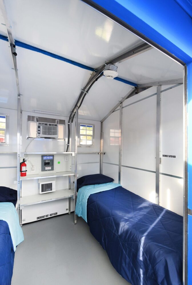 The inside of a tiny home shows two twin beds with blue sheets in a room with white walls and two small windows. There is an air conditioner/heater unit and a fire extinguisher. 