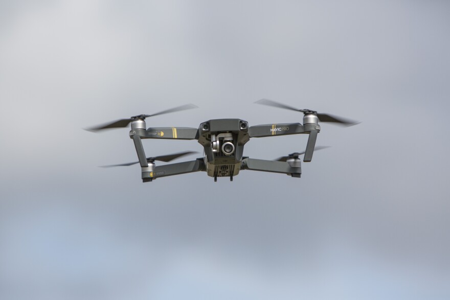 A dark grey drone with four propellers and a camera hovers in mid-air.