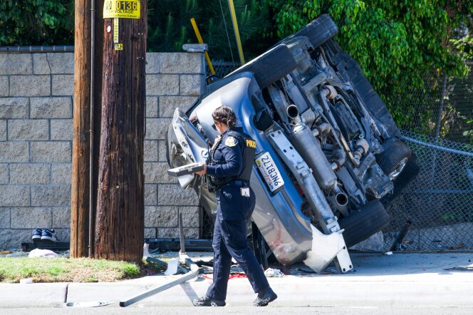 A uniformed police officer walks past a silver sedan that is on it's side, resting against a hollow block fence. There is a wooden electrical pole to the left of the vehicle.