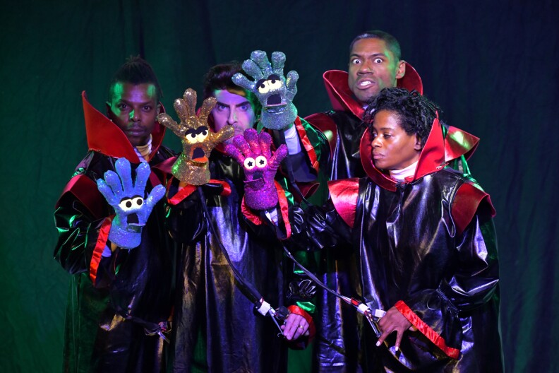 A diverse group of people stand wearing black costumes with red collars. Each of them is holding up a sparkly glove with cartoon eyes and face.