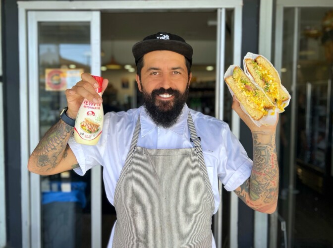 A photograph of a chef with medium dark skin wearing a white short-sleeved shirt and a striped apron holding up a squeeze bottle of mayo in one hand, and in the other hand, he has a large sandwich, cut in two so you can see the insides.