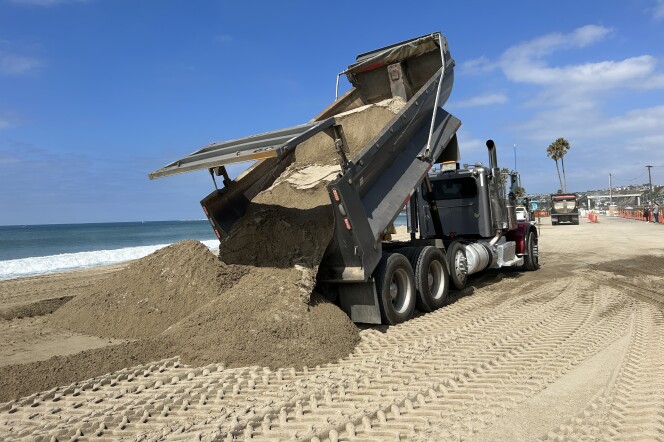 A truck spills a load of sand onto the beach on a day bright with sunshine. Orange work cones and another truck are visible in the background, underscoring the massive job underway.  