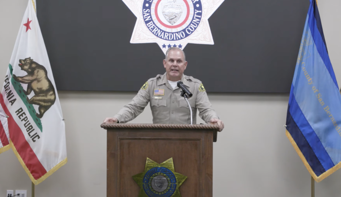 San Bernardino County Sheriff Shannon Dicus addresses a press conference from a podium. He wears a khaki uniform. 