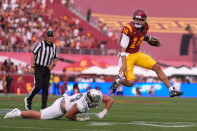 A football player in a cardinal and gold "SC" uniform, number 14, leaps through the air holding the ball while a defender in a white uniform and silver helmet dives at the turf below him.