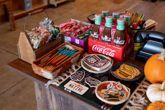 Assorted items on a desk, including stickers for sale that read "Outpost Books' and "Historic Route 66." The table also includes replica classic Coca-Cola bottles, assorted orange and white pumpkins and pencils.