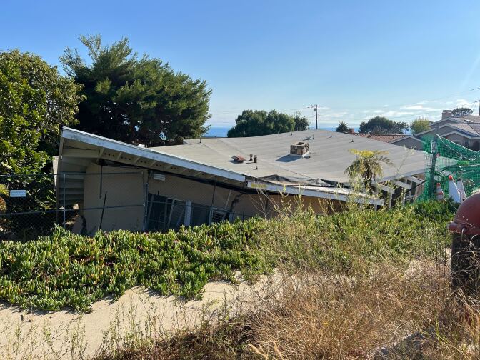 The roof of a home is buckled and jutting up at odd angles. It is surrounded by fencing to keep people from entering.