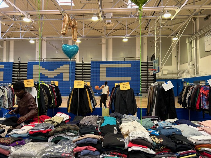 A person with medium-dark skin tone examines a table of shirts. In the background, another person can be seen playing basketball. 