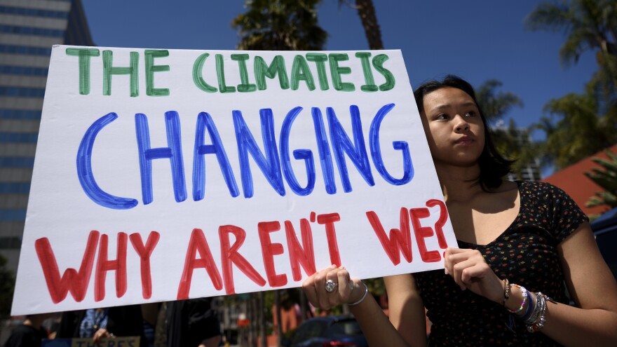 A brown skinned woman wearing a dark colored shirt--- holds up a sign that says "The climate is changing. Why aren't we?"