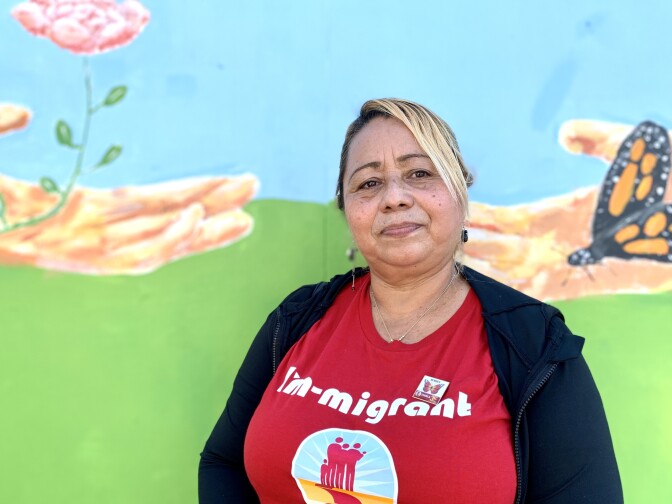 A blonde 56-year-old Latina woman wearing a red "I'm-migrant" shirt stands in front of a mural of two outspread hands, one holding a flower, the other a monarch butterfly.