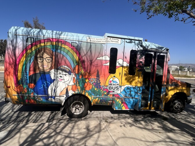A colorful half-size passenger bus is parked next to a sidewalk. It has the image of a sunset, a blue ocean, and a woman wearing glasses carrying a white dog with a hat that says "WINSTON."