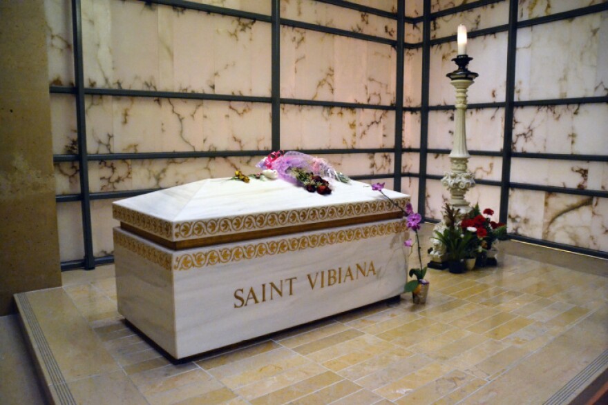 A raised stone container is shaped like a coffin. Saint Vibiana is etched into the front and a decorative trim is around top of box and bottom of lid.