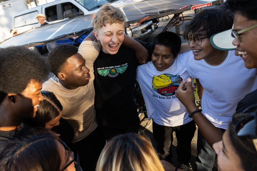 A group of diverse teens huddles in a circle holding wrapping their arms around each other while smiling ecstatically.