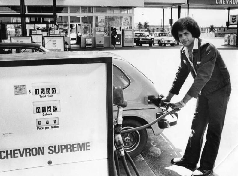 A man in a dark jacket and pants with an afro fills up a hatchback car.