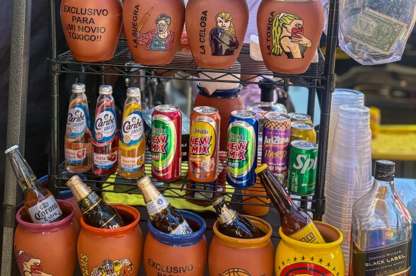 Painted cantaritos are displayed on shelving, with signs that read “RAIDERS,” “LA SUEGRA,” and “LOS ANGELES LAKERS.” The shelves also hold a variety of imported alcoholic drinks, plastic cups, and a transparent cup with a dollar bill inside.
