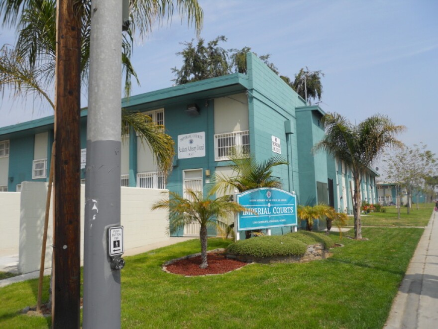 A teal colored apartment building with white trim. It has a sign in front of it that reads Imperial Courts. Palm trees surround the building.