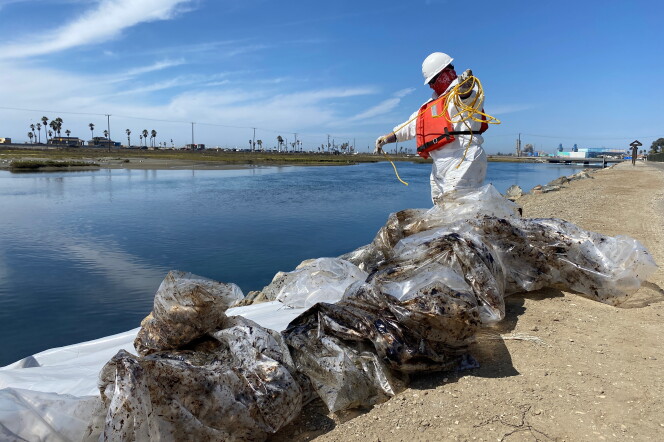 Plastic bags full of oil debris sit on the shore of a channel with a worker in white coveralls and an orange life preserver coils yellow rope.