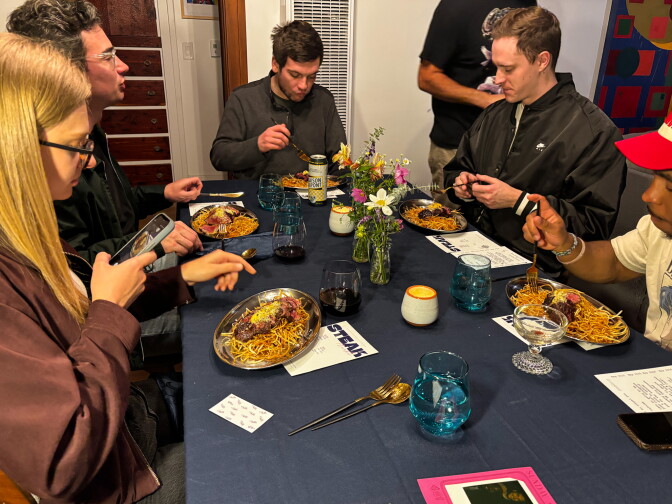 Six guests sit around a navy tablecloth-covered dining table eating hanger steak frites from silver oval plates, with fresh flowers, blue glassware, wine, and Steak Freaks menus visible on the table, inside a warmly lit living room.