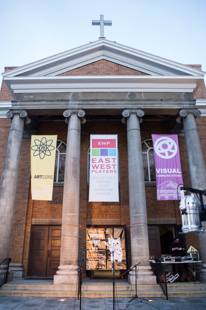 A DJ stand in front of a turntable setup in front of a church featuring four ionic Greek columns. Three banners hang down that read "East West Players," "Visual Communications" and "ArtCORE."