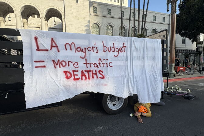 A man with an orange helmet lays on the road behind a truck with a white rose in his right hand. The truck is draped with a white sheet and the following painted on it in red: LA Mayor's budget = more traffic DEATHS.