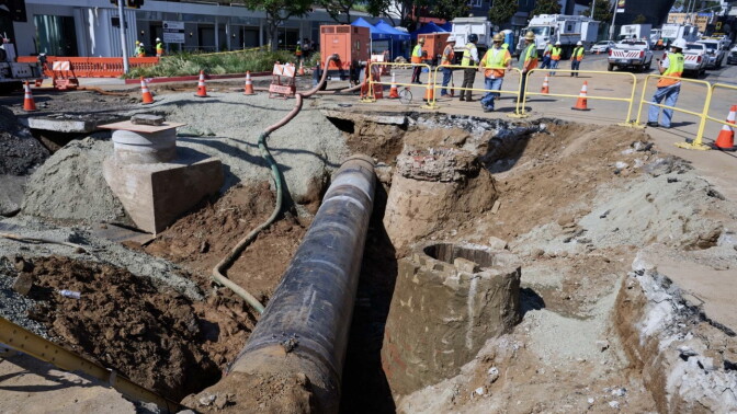 A section of a city street has been dug up, creating a deep trench with an exposed pipe running down the middle. 