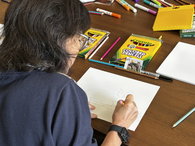 A woman sketches a cat on paper, surrounded by Crayola colored pencils and markers on a wooden table.