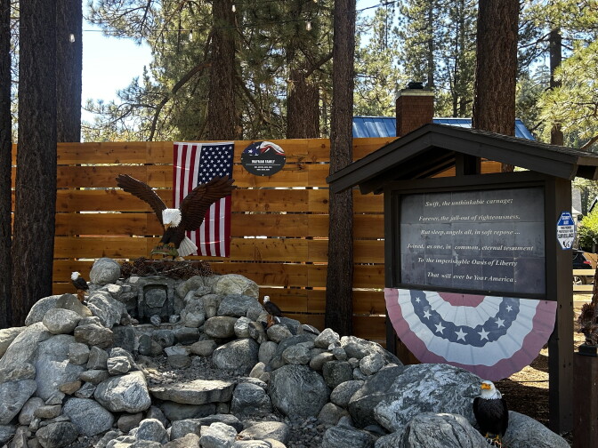 A memorial display with an eagle statue, American flag, and a sign bearing a tribute poem sits among rocks near a wooden fence.