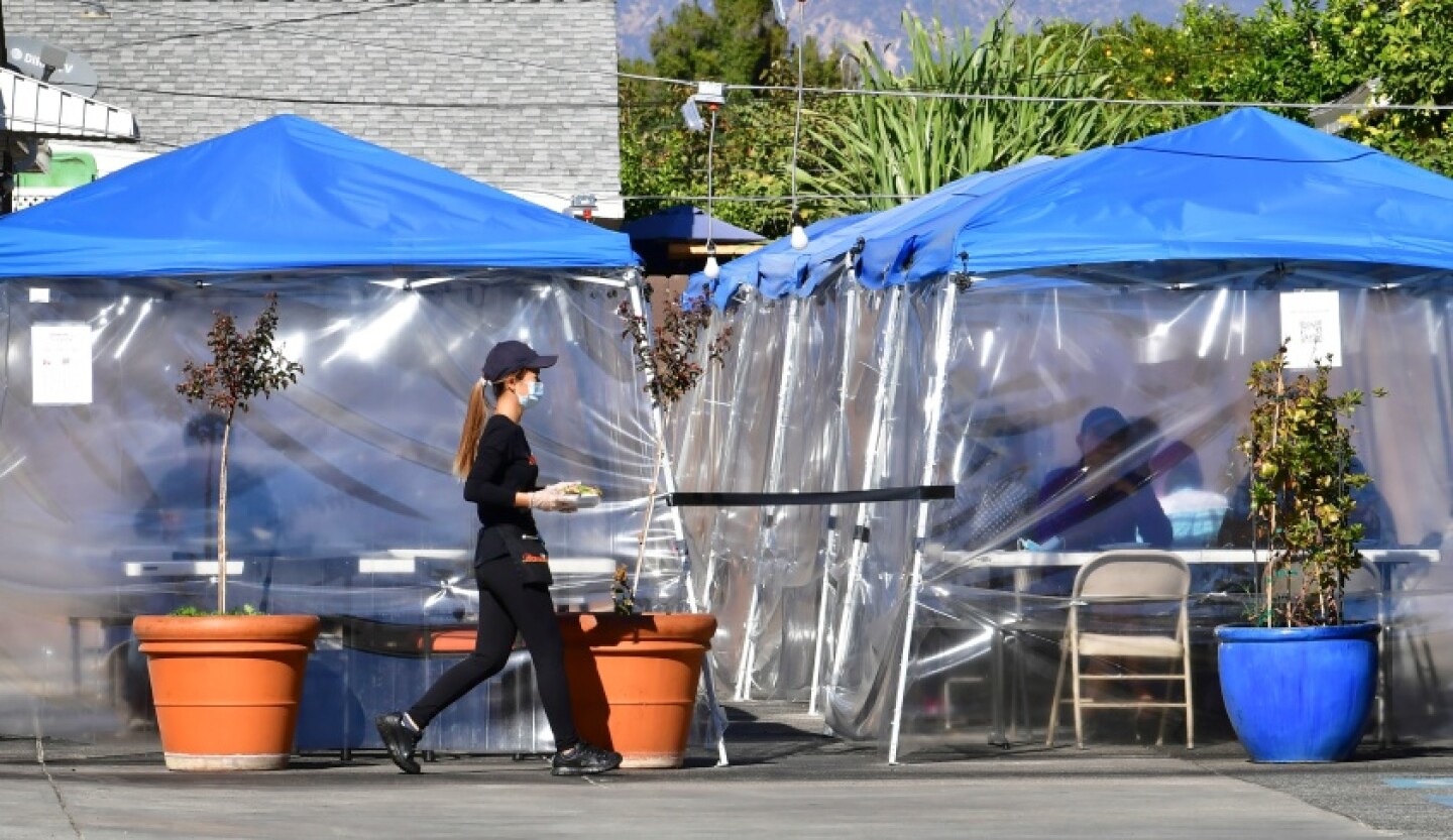 An image of a restaurant worker in dark clothing walking toward diners who are eating outdoors in plastic covered tents.