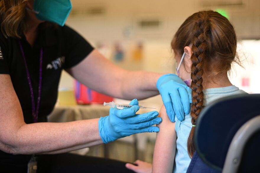 A person with a face mask and blue gloves on administers a shot to a child's arm. The child is facing the opposite of the camera and has a braid.