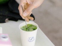 A light skinned hand hold a metal spoon as it mixes a green liquid in a paper white cup
