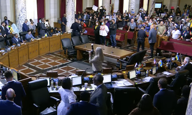A crowd of people stand inside a large city chamber as some people stand around or sit at a dais.