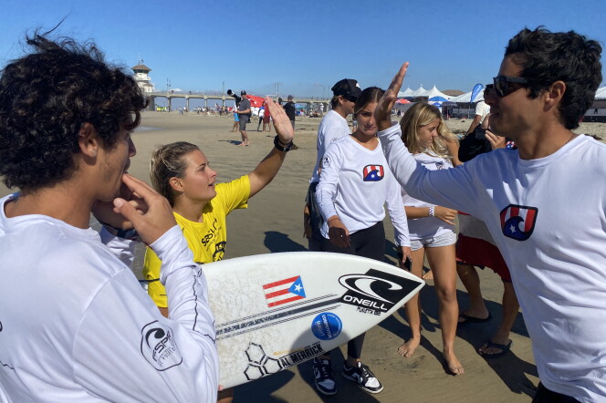 A woman with blond hair and wearing a yellow jersey holds a surfboard with a Puerto Rican flag on it while high-giving with a man in a white t-shirt. Other people gather around. 