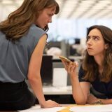 Carey Mulligan sits left of the frame on top of a long wood table. Her back is towards the viewer as she leans slightly to her right looking across her shoulder at Zoe Kazan who sits right of her. Mulligan's face can be seen in profile, her expression serious and tense. She wears a grey tank tucked into black pants, her right arm braced on the table to help her lean. Zoe Kazan leans over the table to Mulligan's right. Kazan faces the viewers as she looks at Mulligan, her body angled left. Her left arm rests in front of her on the edge of the table while her right arm is propped up on her elbow as she holds a gold colored phone in her hand. It is facing up as if on speaker phone with someone. Kazan wears a navy blue t-shirt. She looks up at Mulligan only with her eyes, she expression is also tense. They women seem to be sharing a knowing look with each other as they listen to whatever is being said on the phone. Behind them are empty rows of black desktop computers and white walls of a newsroom. 