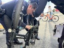 Police officers examine a folded electric bike's gears in a parking lot as another writes on a clipboard.