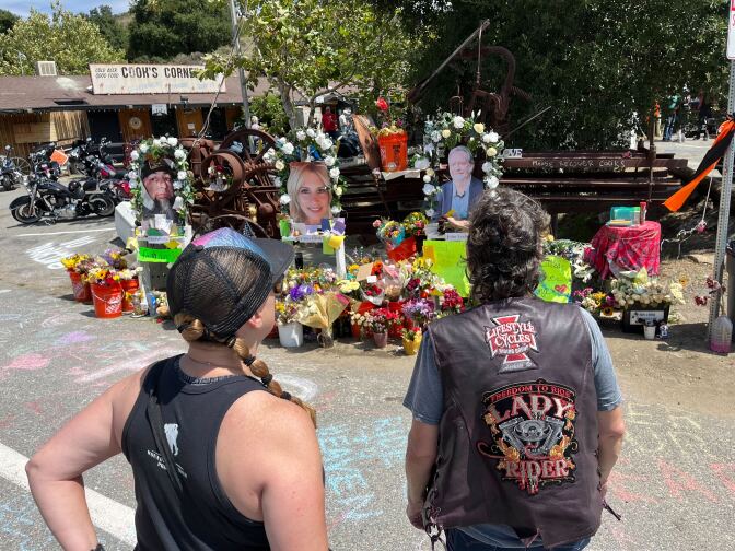 Two people -- including one wearing a motorcycle vest -- pause in front of a makeshift memorial decorated with buckets and pots filled with flowers, signs, notes and three life-sized photos of the victims. 