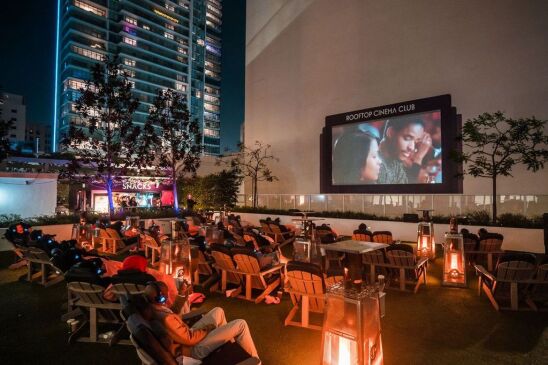 The image is people seated at the Rooftop Cinema Club enjoying there movie. The people are outside at night, all seated in cozy wooden chairs in a nicely dim surrounding. Everyone is also wearing provided over the ear headphones to actively listen and enjoy the movie instead of a usual sound system. 