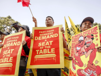 Three protesters stand side by side. Two are holding large posters that read Fast Food Workers Demand A Seat At the Table.