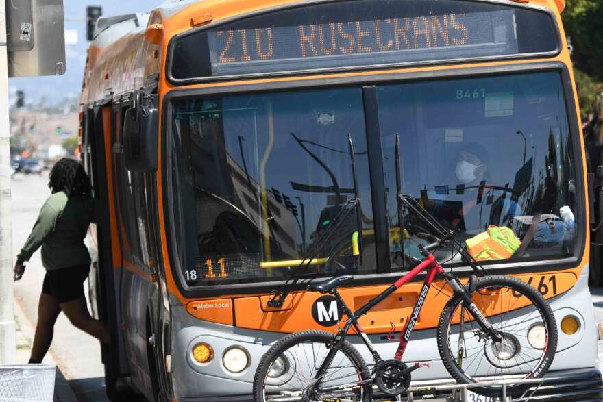 An image of a woman steps out to the curb from the rear door of an orange L.A. Metro bus as the driver, wearing a face mask, watches.