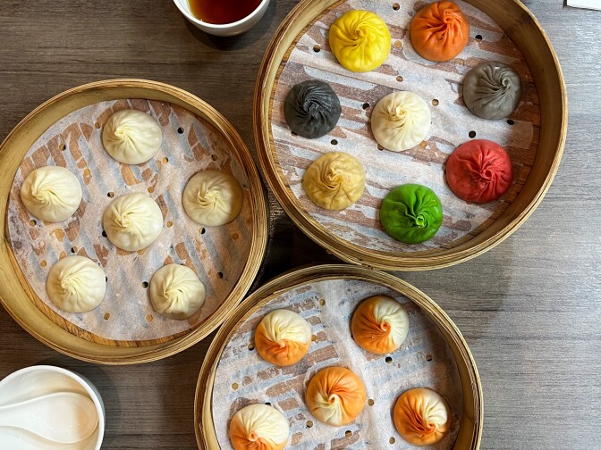 A photograph of three sets of dumplings in bamboo steamer baskets, each a different color, sitting on a wooden table. The bottom group of dumplings is orange and white. The center group is white. The upper right basket contains dumplings that are green, red, grey, back, yellow, orange and white. 